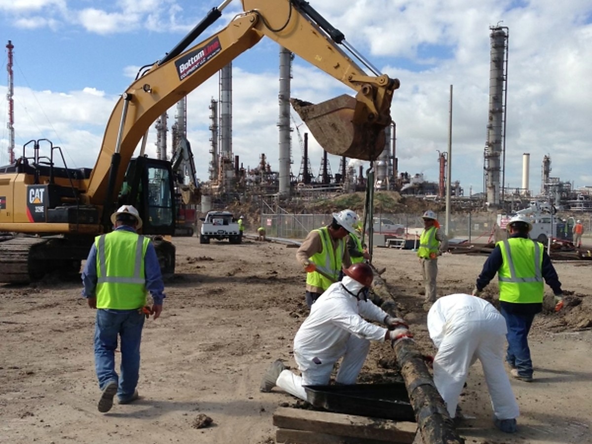 Crew in protective suits and hi-vis removing coating from pipe at a refinery site