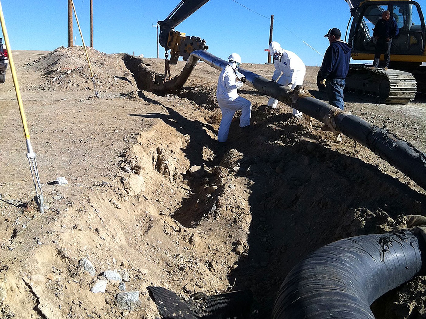 Crew in protective suits and respirators removing coated pipe from an open trench