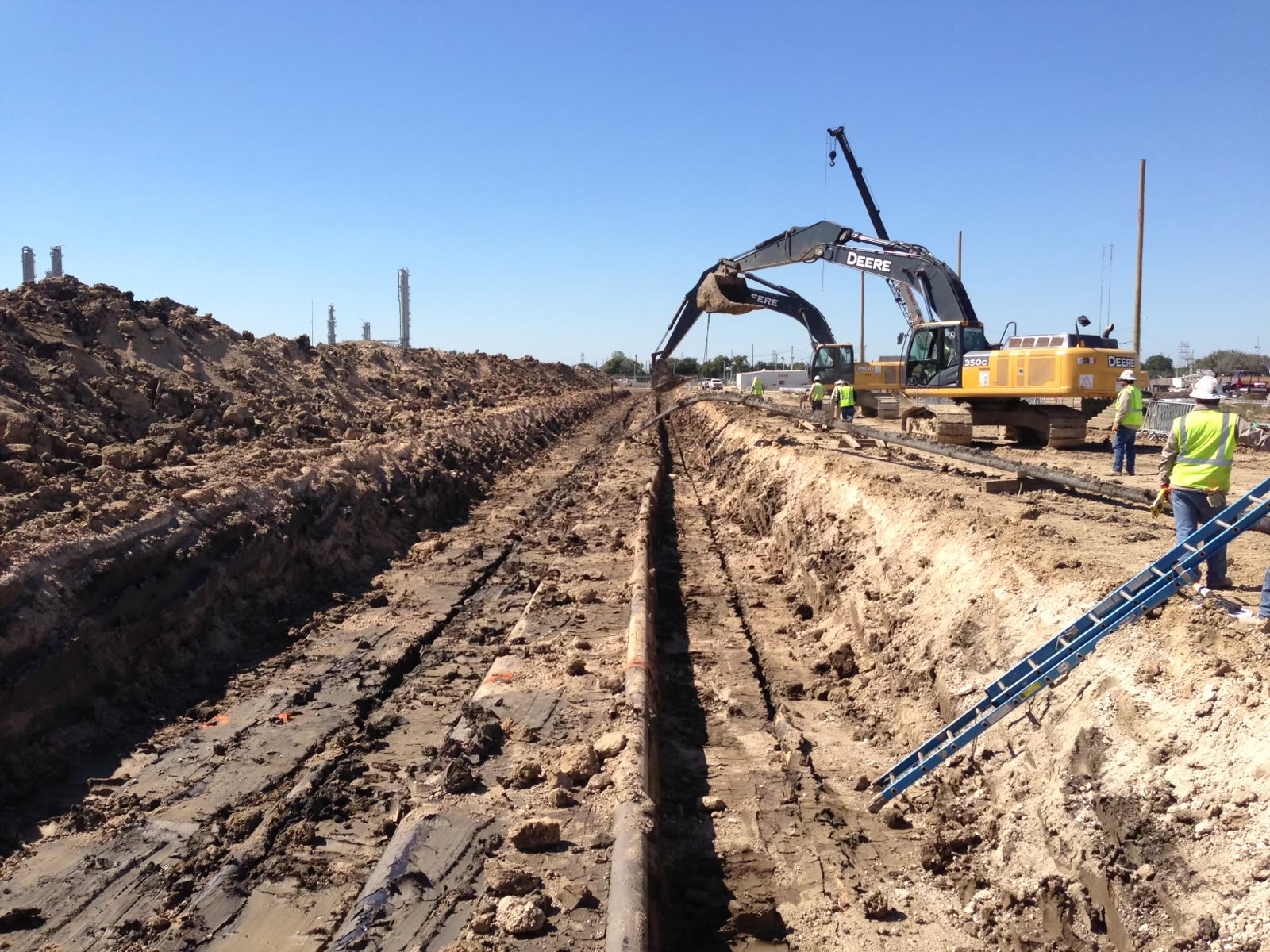 Open trench with the exposed line running to the horizon, excavator and crew working the right-of-way