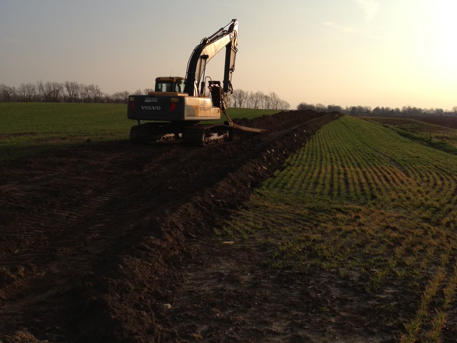 Excavator working the right-of-way at sunset, spoil windrowed alongside the trench