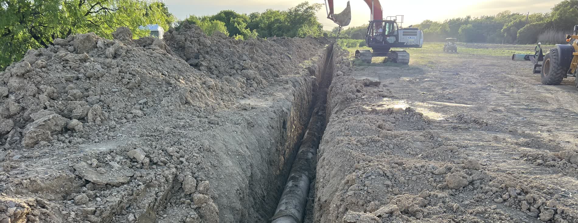 Open trench with exposed line pipe and an excavator working the right-of-way at dusk