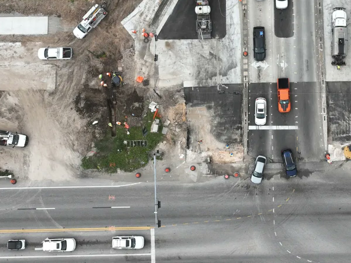Aerial view of a pipe section being excavated from beneath the Highway 146 corridor, with an excavator and crew in the trench and live traffic queued behind cones a few feet away